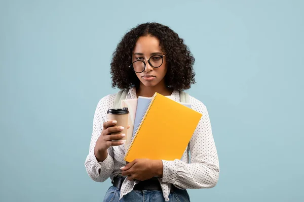 Tired young black female student with notebooks and takeout coffee looking at camera on blue studio background — Fotografia de Stock