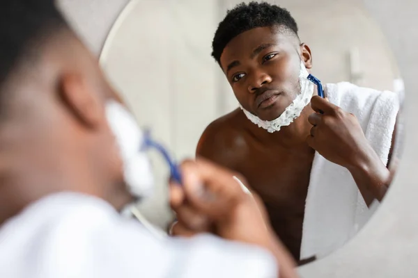 Serious African Male Shaving Face With Razor In Modern Bathroom — Stock fotografie