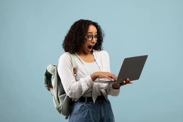 Онлайн-освіта. Емоційна молода чорна жінка з рюкзаком дивиться на екран ноутбука в збудженні на синьому фоні — стокове фото