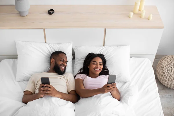 Glad smiling young black female and male, lying on bed, typing on phones, chatting, playing online game — 스톡 사진