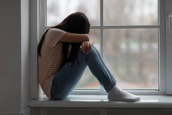 Molesto chica morena de pelo largo sentado en el alféizar de la ventana y llorando —  Fotos de Stock