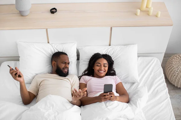 Displeased young african american guy gesturing, spreads hands, look at woman smartphone on bed — 스톡 사진