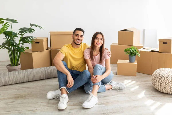 Full length portrait of loving multinational couple sitting on floor among cardboard boxes, hugging and looking at camera