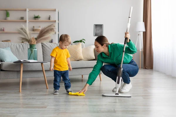 Little kid boy watching her mother mopping the floor after him. Woman wiping the floor with rag next to her baby — Fotografia de Stock