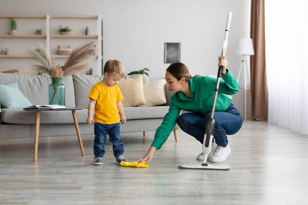 Little kid boy watching her mother mopping the floor after him. Woman wiping the floor with rag next to her baby