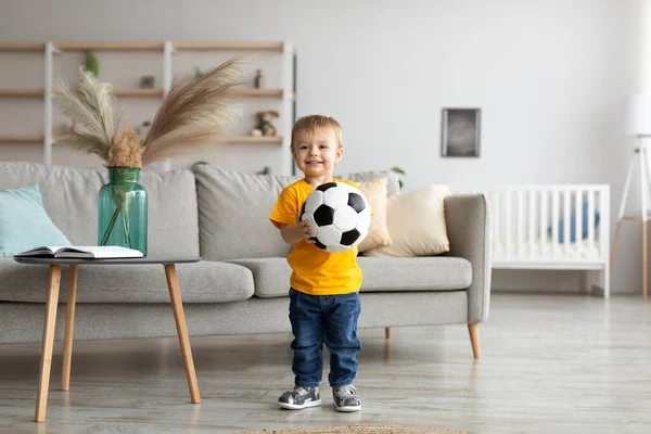 Happy toddler boy football fan posing with soccer ball and smiling at camera, having fun at home in living room — Stockfoto