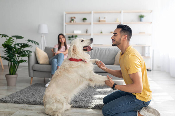 Happy young Arab guy playing with dog, having fun on floor at home, his girlfriend drinking coffee on background