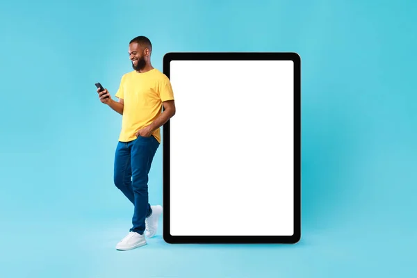 Optimistic young black man leaning on huge tablet computer with blank white screen, using mobile phone, mockup — стоковое фото