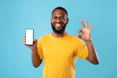 I recommend this new mobile app. Young black guy showing empty smartphone and gesturing okay on blue background, mockup