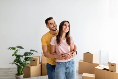 Positive young interracial couple hugging in their apartment among carton boxes on moving day, free space