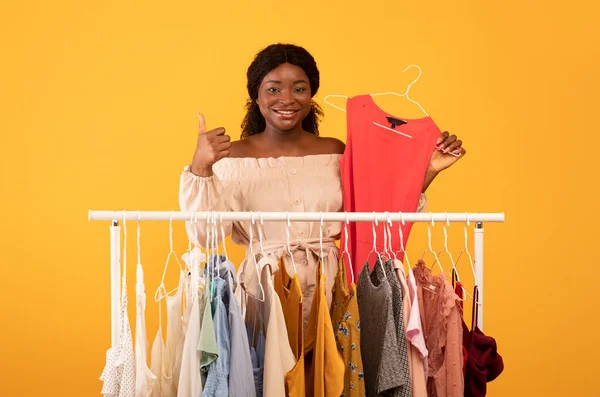 Attractive young black woman holding dress near clothing rack, showing thumb up gesture, recommending new trendy look — Stok fotoğraf