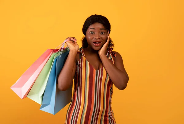 Excited black female with shopping bags touching her face in surprise, cannot believe huge sale on orange background — Stockfoto