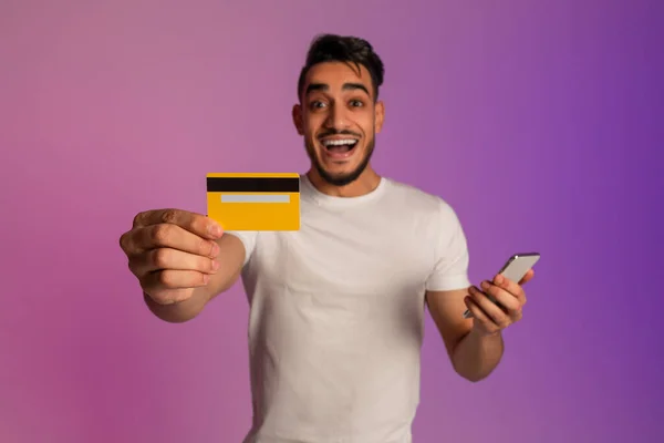 Online shopping app. Joyful millennial Arab man holding cellphone, showing credit card in neon light, selective focus — Zdjęcie stockowe