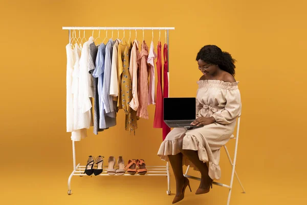 Beautiful young black woman sitting near clothes rack with laptop, offering mockup for web store design on screen — Foto Stock