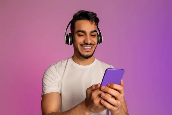 Portrait of cool young Arab man in wireless headphones using smartphone in neon light — Fotografia de Stock