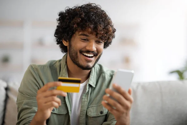 Happy guy sitting on couch with credit card and cellphone — стоковое фото