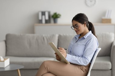 Professional psychologist writing in clipboard, sitting on chair at office, offering professional counseling services