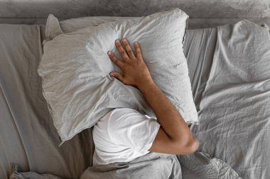 Stressed black man covering ears and head lying in bed