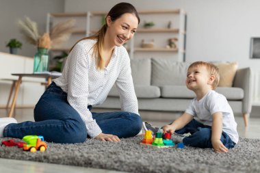 Happy mom playing with toddler son stacking and sorting wooden toy, spending time with kid at home