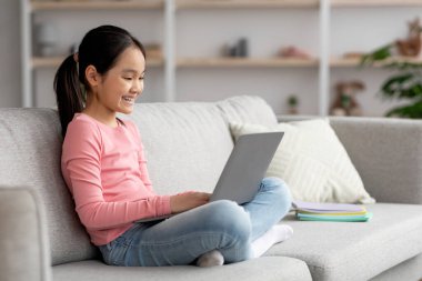 Cheerful chinese child using laptop, chilling at home, side view