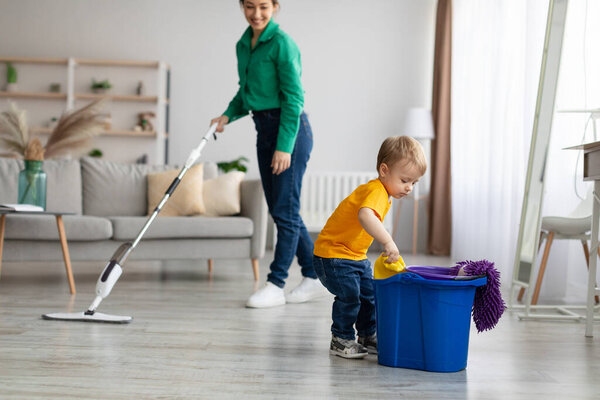 Little helper. Adorable toddler boy helping mom cleaning at home, kid getting rag out of bucket while mom mopping floor