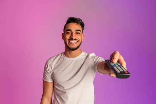 Positive young Arab guy watching TV, switching channels with remote control in neon light — Fotografia de Stock