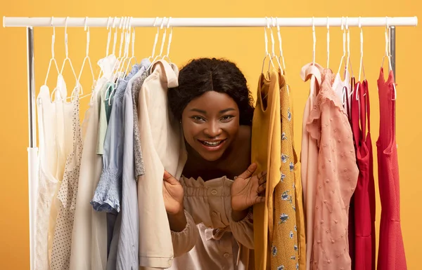 Cheerful millennial black female peeking out between clothes hanging in rail, smiling at camera over orange background — Stok fotoğraf