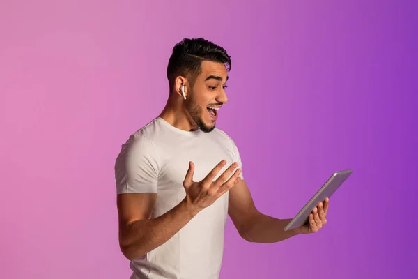 Excited Arab guy in earphones looking at tablet pc, shouting OMG, overjoyed about huge win or online sale in neon light — Stockfoto