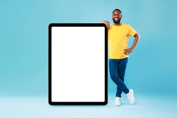 Cheerful young black guy leaning on giant tablet pc, demonstrating blank screen on blue background, mockup — ストック写真