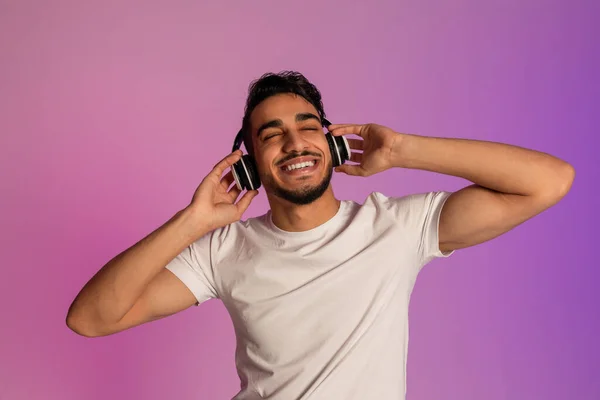 Peaceful young Arab guy listening to music in wireless headset, closing eyes and relaxing in neon light — Zdjęcie stockowe