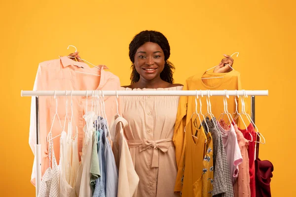 Shopping and fashion. Beautiful young black woman choosing outfits near clothing rack on orange studio background — Stok Foto