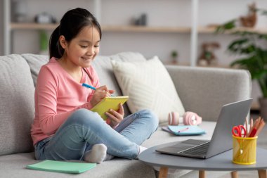 Smart asian kid studying at home, taking notes, using laptop