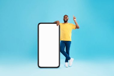 Handsome black guy offering mockup for new mobile app on giant smartphone screen, pointing upwards on blue background