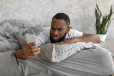 Kafası karışmış siyah adam yatakta telefona bakıyor.