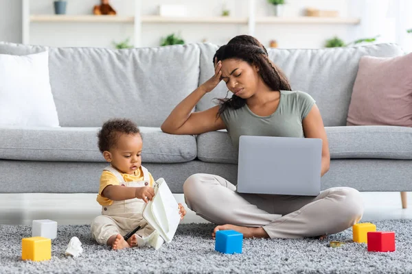 Stressed Black Woman Trying To Work On Laptop With Little Baby Around — стоковое фото