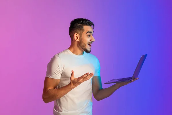 Handsome young Arab guy looking at laptop screen, excited about online lottery win or success in neon light — Stock Fotó
