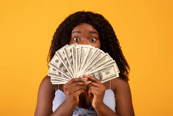 Shocked African American woman covering her face with money, showing shopping budget on orange background — Stok fotoğraf