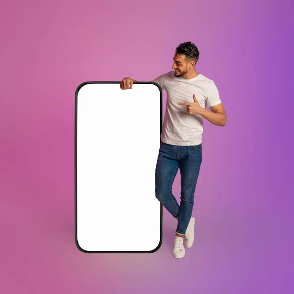 Happy young Arab man leaning on huge cellphone with empty white screen, gesturing thumb up, recommending new app, mockup — ストック写真