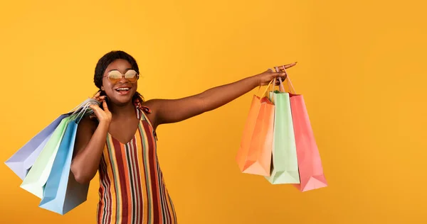 Pretty young black woman in dress and sunglasses holding gift bags, pointing at blank space over orange background — Stockfoto