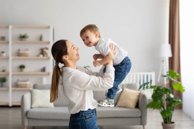 Emotional young caucasian mother raising laughing toddler son, having fun and playing together in light living room