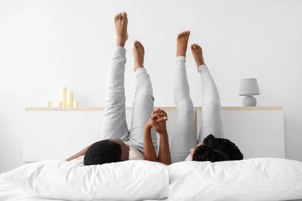 Young african american guy and lady in love lie on white bed in domestic clothes hold hands, raise legs up — Foto Stock