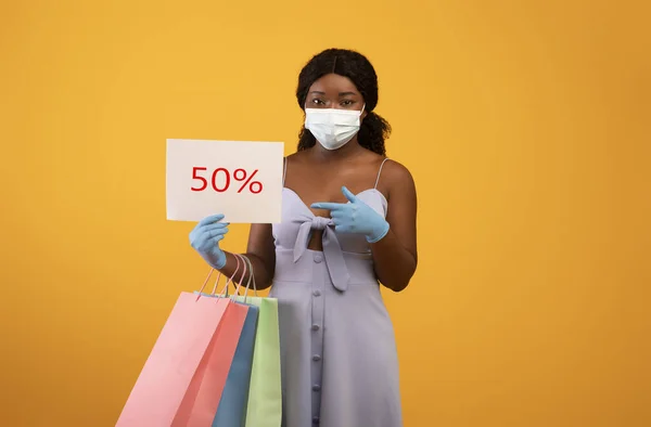 African American lady in surgical mask and rubber gloves holding FIFTY PERCENT OFF sign and shopping bags — Stock fotografie