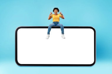 Positive millennial black woman showing thumb up gesture, sitting on top of huge smartphone over blue background, mockup