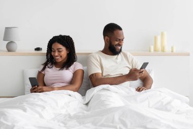Glad young black female ignores her husband sitting on bed and look at phone in bedroom interior, copy space