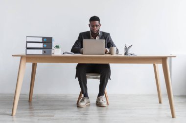 Full length of focused young black man working online with laptop computer, sitting at desk against white studio wall