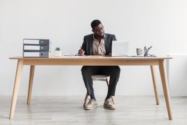 Serious African American businessman in suit writing in notebook and using laptop computer, sitting at desk