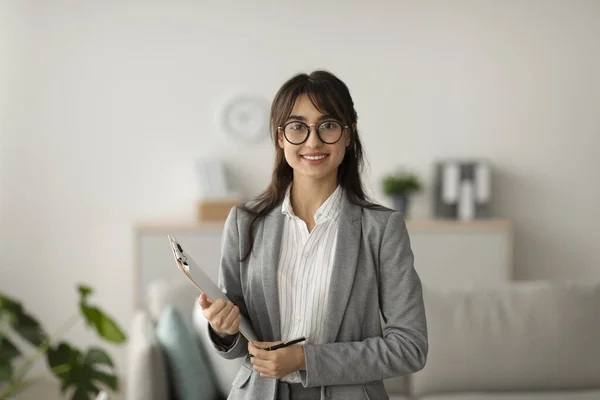 Portrait of cheerful female psychologist posing with clipboard, looking and smiling at camera, working in modern office — 图库照片
