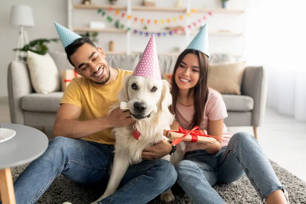 Millennial multiracial couple in party hats holding gift box, celebrating pet dogs birthday at home — стоковое фото