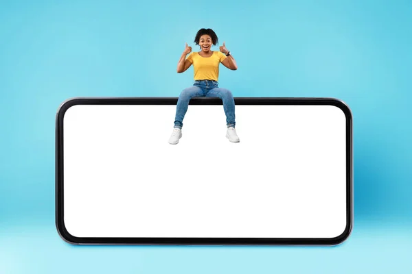 Positive millennial black woman showing thumb up gesture, sitting on top of huge smartphone over blue background, mockup — Stok fotoğraf