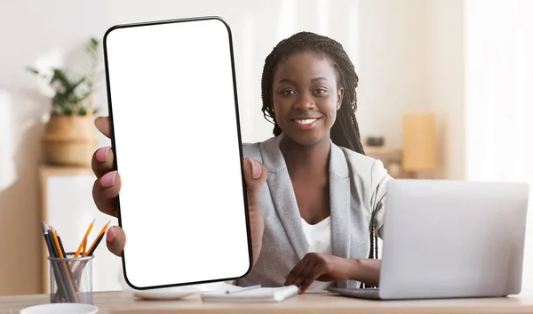 Cheerful black businesswoman showing smartphone with big blank screen at modern office, mockup for mobile app or website — Fotografia de Stock